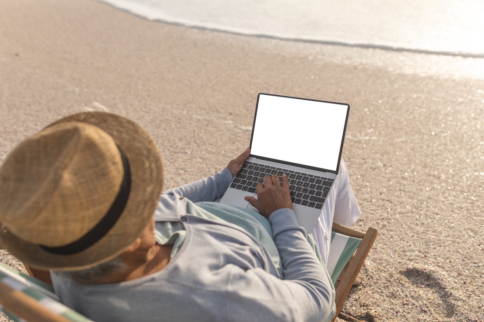 Biracial senior woman wearing hat using blank screen laptop with copy space at beach during sunset. lifestyle and telecommunications.